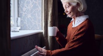 An old lady seated by her window sipping her tea while touching the heater